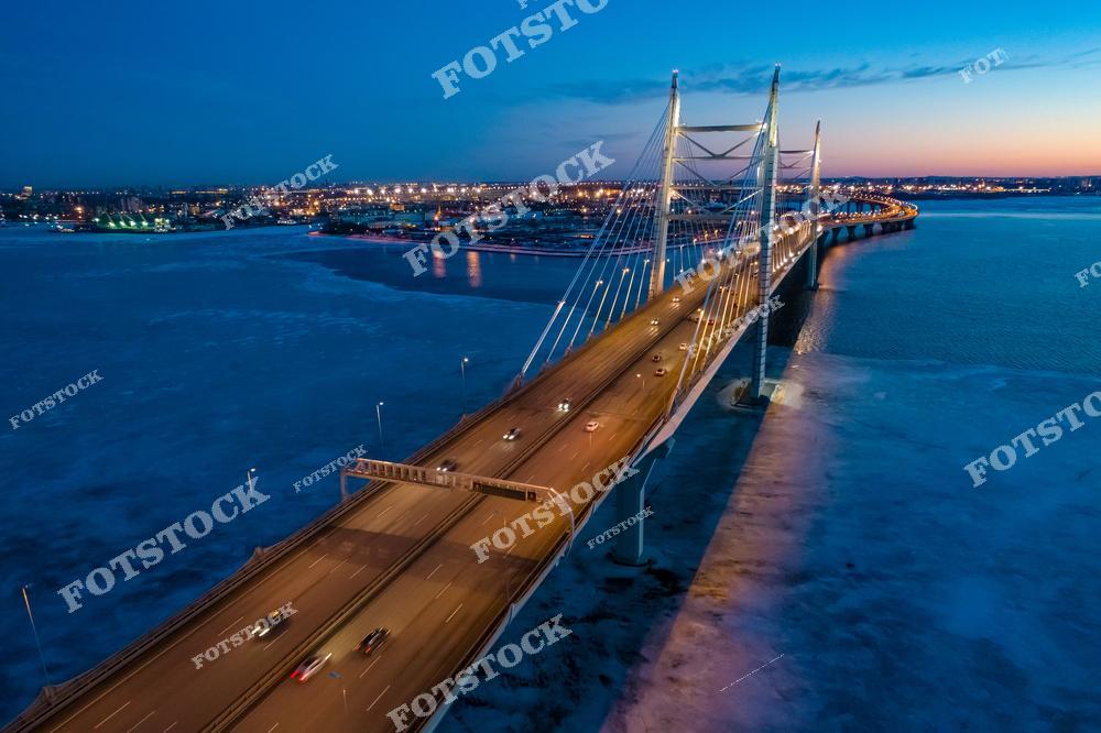 На фотографии изображен Большой Обуховский мост, известный как Вантовый мост, в вечернее время. Мост соединяет берега реки Невы, часть которой покрыта льдом, что создает зимний пейзаж. Яркие огни вдоль моста и городская иллюминация вдалеке подчеркивают современность и динамику Петербурга. Снимок с высоты демонстрирует величие моста и его значение как важной транспортной артерии города.